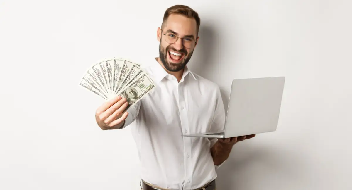 The image shows a cheerful man with glasses, a beard, and a white shirt, holding a laptop in one hand and fanned-out US dollar bills in the other. He appears excited or happy, after selling his old laptop to MS Scrap Buyers Hyderabad.