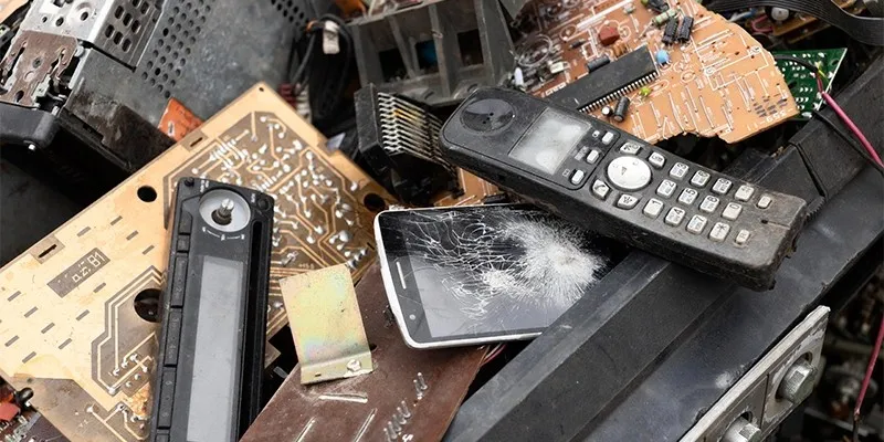 A pile of discarded electronic devices and appliances, including old computer monitors, a keyboard, a printer, a dishwasher, a microwave, and various other e-waste items, is overgrown with greenery, including small trees and plants. The recycling symbol prominently displayed in the center highlights the environmental impact of e-waste in Hyderabad and emphasizes the importance of proper disposal and recycling practices to mitigate harm to the environment.