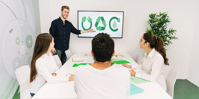 A team of professionals seated in a modern, white-walled meeting room with a large wall graphic are engaged in a discussion about sustainability. One person, standing and pointing to a screen displaying three green recycling symbols, is presenting strategies for a sustainable future. The image emphasizes collaboration and strategic planning in promoting environmental sustainability.